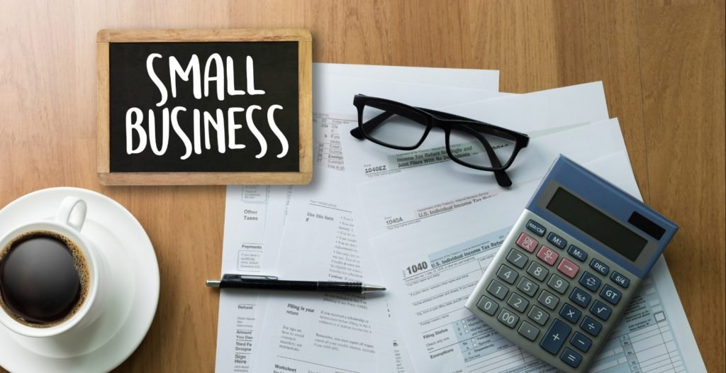 A wooden table with a small business black board, papers, caluculator, eyeglasses, ball pen and coffee placed on top.